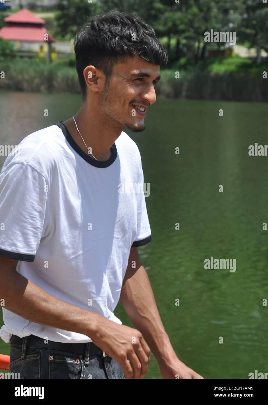 Porträtaufnahme eines jungen indischen Mannes, der neben dem See mit einem weißen T-Shirt steht und lächelt Stockfoto