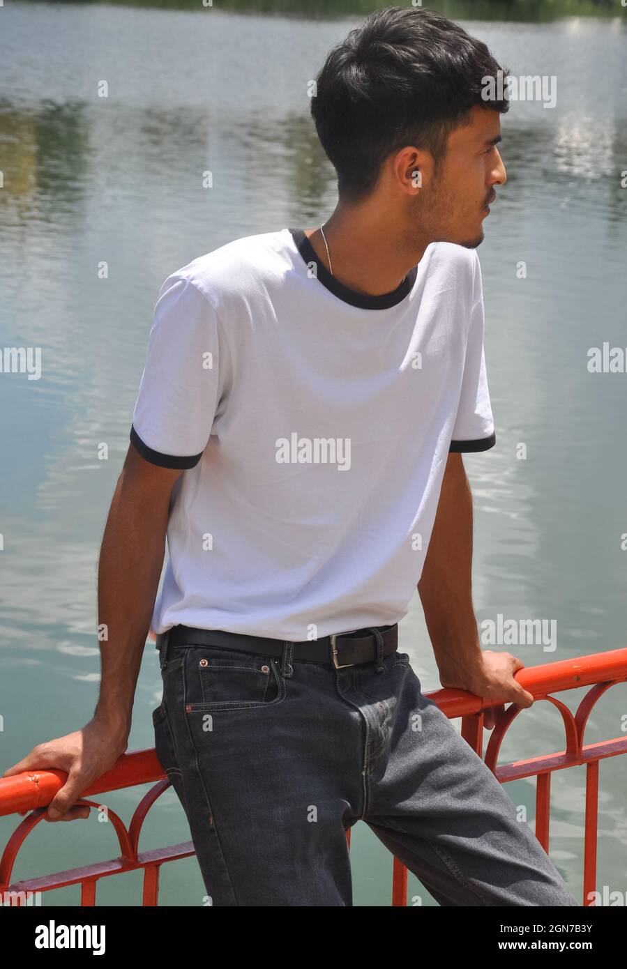 Ein gut aussehender indischer junger Mann, der weißes T-Shirt und schwarze Jeans trägt und auf einer Sicherheitsbarriere am See sitzt und zur Seite schaut Stockfoto