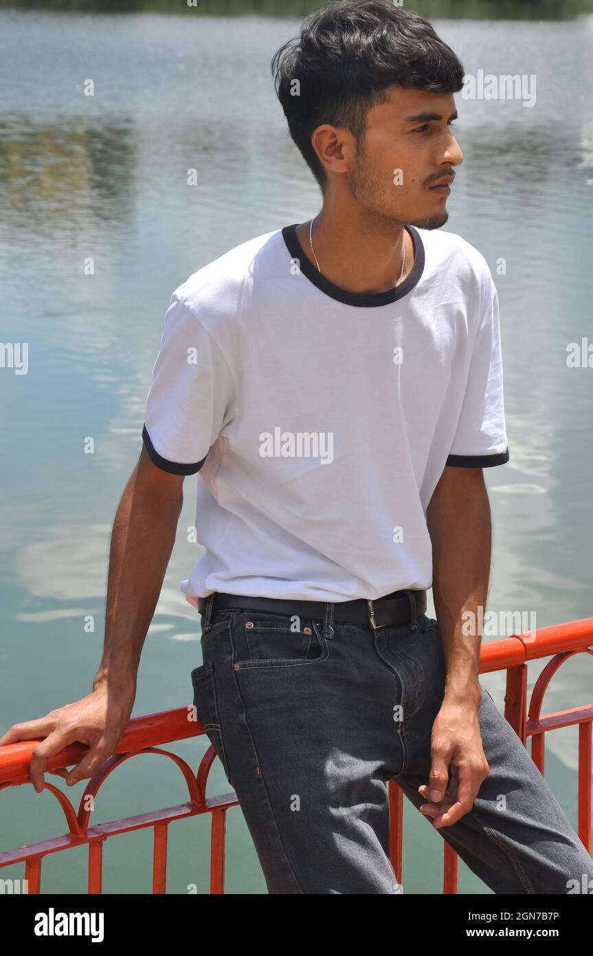 Ein gut aussehender indischer junger Mann, der weißes T-Shirt und schwarze Jeans trägt und auf einer Sicherheitsbarriere am See sitzt und zur Seite schaut Stockfoto