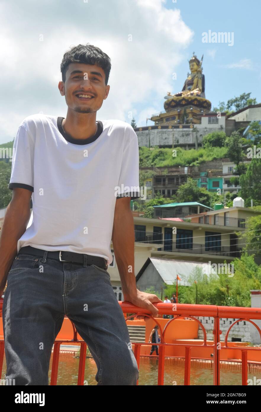 Ein hübscher Typ, der lächelt und auf einer Sicherheitsbarriere am See und in der wunderschönen hügeligen Gegend von Tso-Pema sitzt und im Hintergrund die Statue von Padmasambhava (Guru Rinpoche) sieht Stockfoto