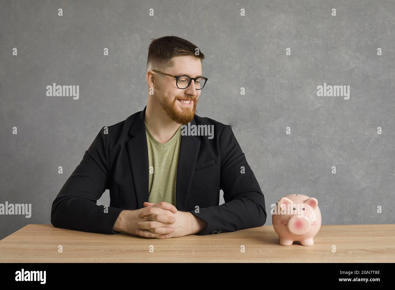 Fröhlicher, gutaussehender Mann, der am Tisch sitzt und mit einem Lächeln auf dem Gesicht auf das Sparschwein blickt. Stockfoto
