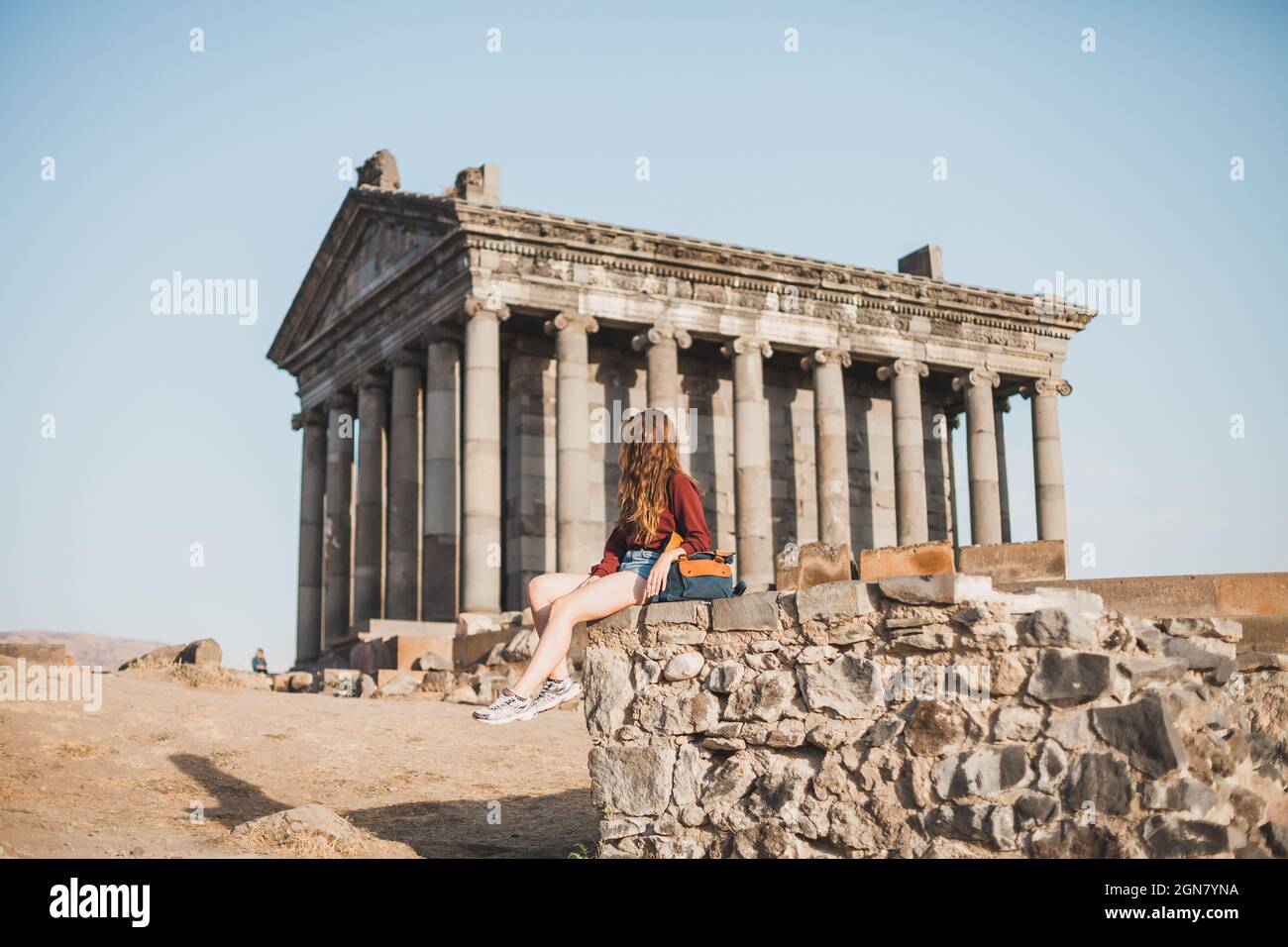 Junge Frau, die in der Nähe des alten Steingebäudes mit Säulen in Armenien sitzt. Tourismus in Armenien. Reisende, die ein altes Architekturgebäude betrachten. Stockfoto
