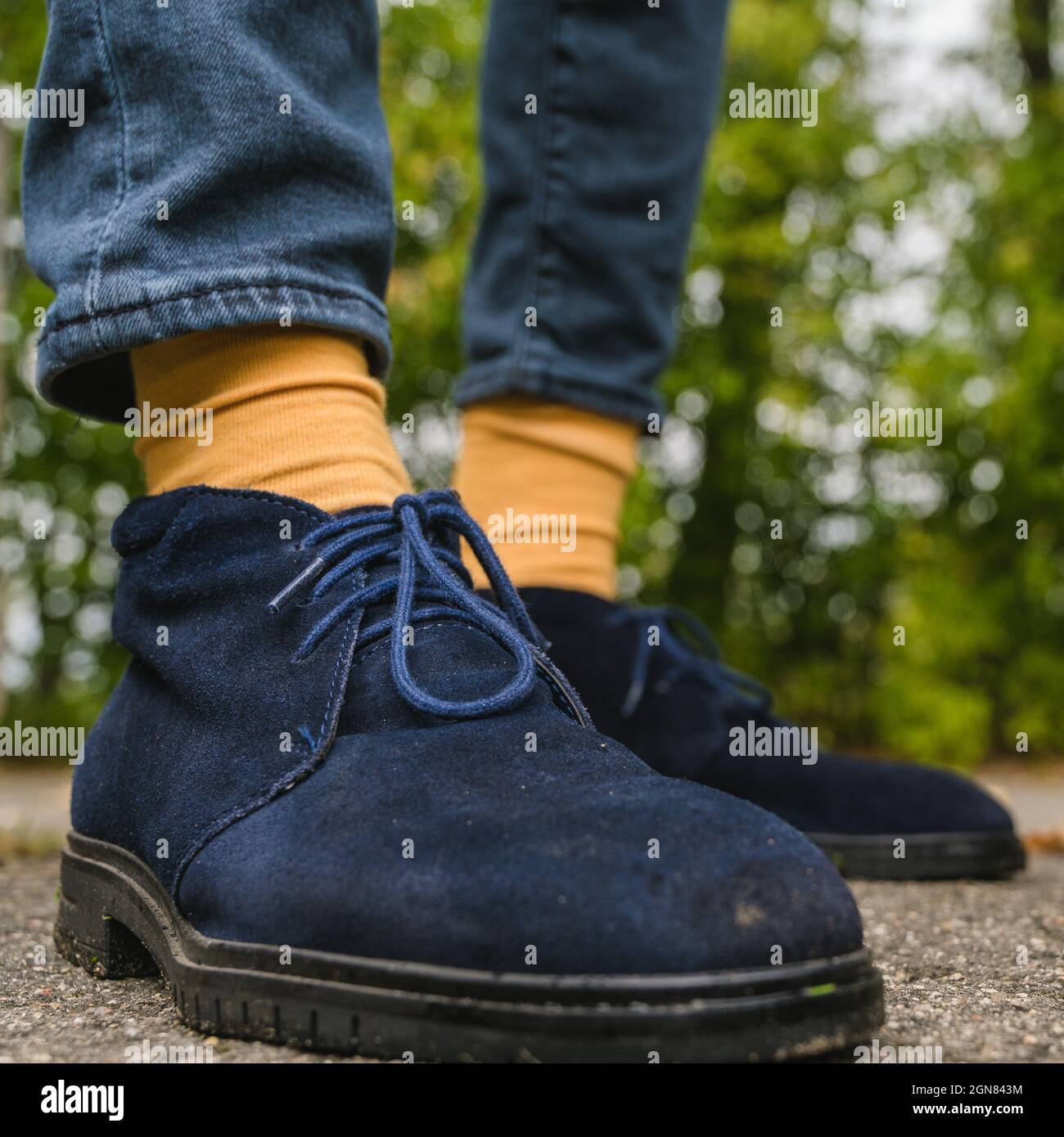 Nahaufnahme der Beine in blauen Wildlederschuhen und gelben Socken. Asphalt mit abgefallenen Blättern. Herbstmode für Männer. Stockfoto