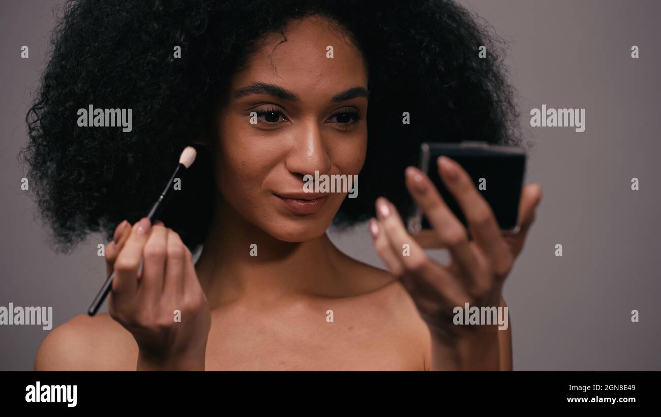 Junge afroamerikanische Frau mit kosmetischen Pinsel Blick auf Spiegel isoliert auf grau Stockfoto