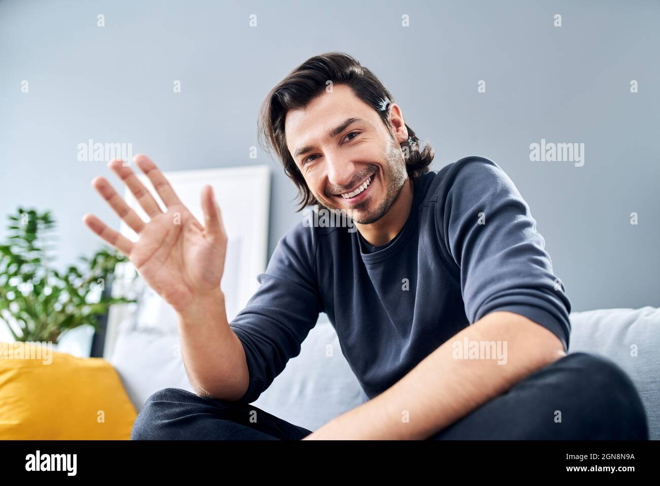 Lächelnder Mann winkt mit der Hand, während er zu Hause auf dem Sofa sitzt Stockfoto