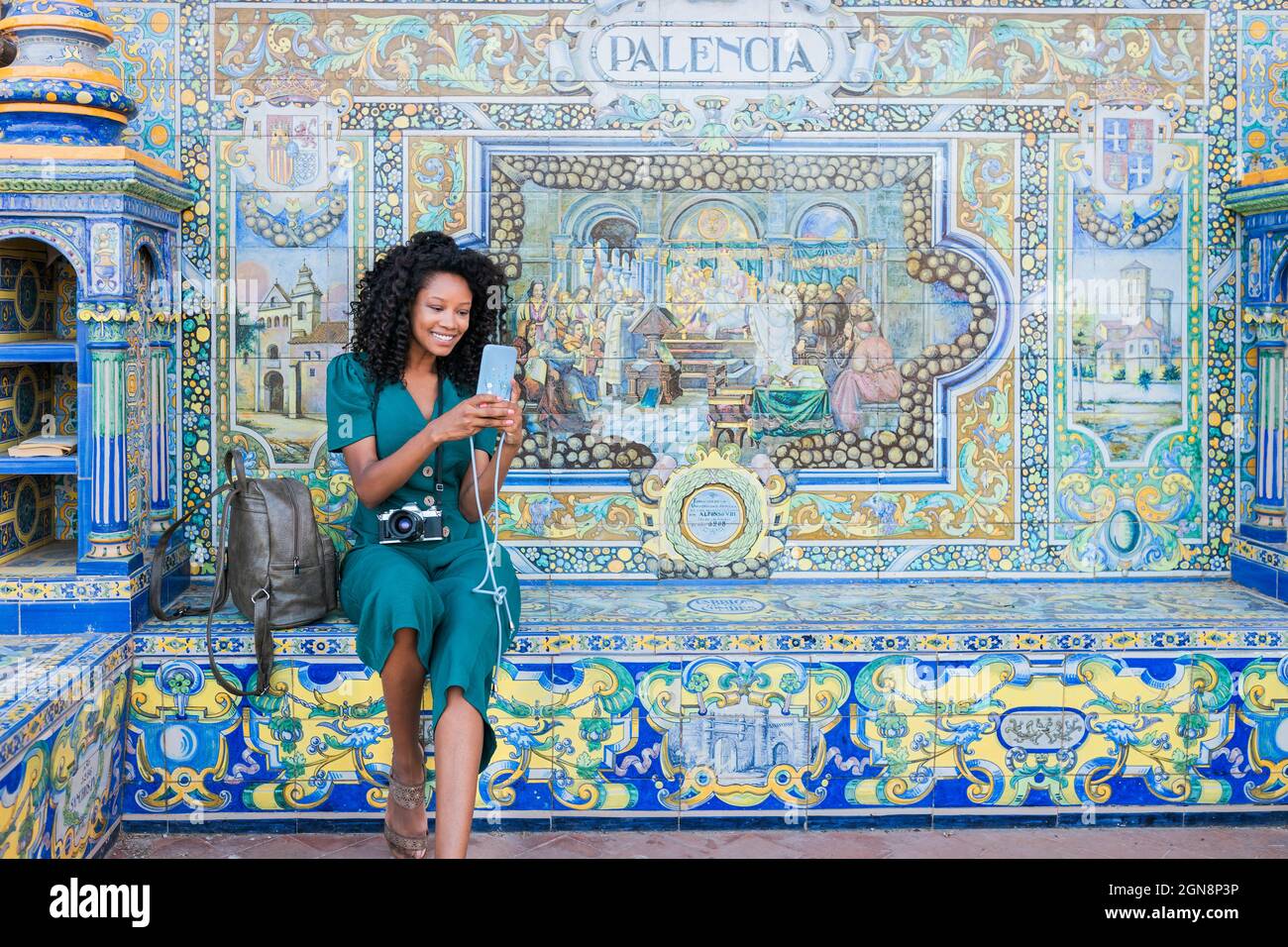 Lächelnde junge Frau, die Smartphone benutzt, während sie vor dem Mosaik auf der Plaza De Espana, Sevilla, Spanien sitzt Stockfoto