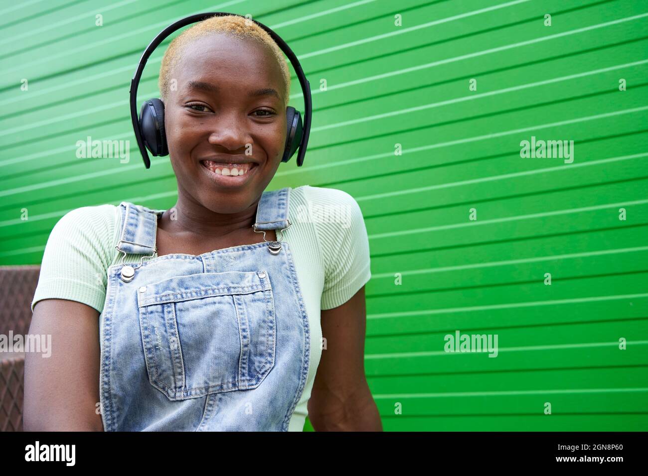 Glückliche Frau mit kurzen blonden Haaren, die Musik über kabellose Kopfhörer an der grünen Wand hört Stockfoto
