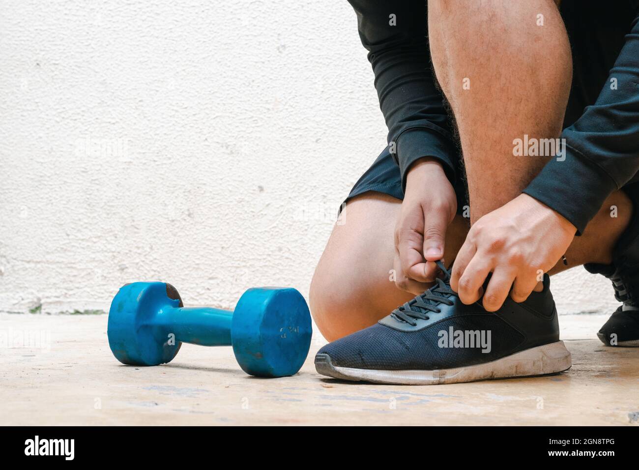 Knieender Mann, der seine Schuhe vor dem Training neben Hantel und Flasche mit blauem Wasser bindet. Training zu Hause, weißer Hintergrund. Fitnesskonzept. Heilen Stockfoto Knieender Mann, der seine Schuhe vor dem Training neben Hantel und Flasche mit blauem Wasser bindet. Training zu Hause, weißer Hintergrund. Fitnesskonzept. Heilen Stockfoto