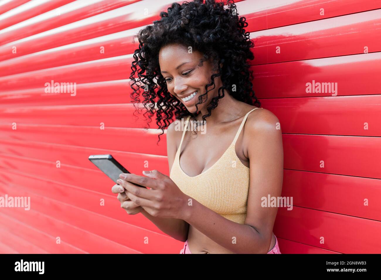 Glückliche Afro-Frau, die über das Mobiltelefon im Netz surft, während sie sich auf den roten Verschluss stützt Stockfoto