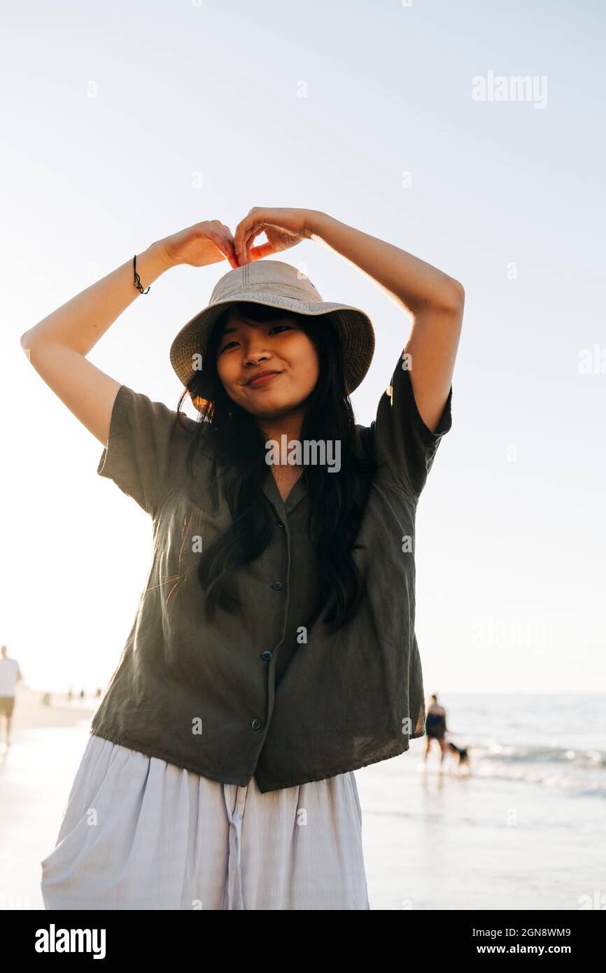 Lächelnde junge Frau mit Hut gestikuliert am Strand Stockfoto