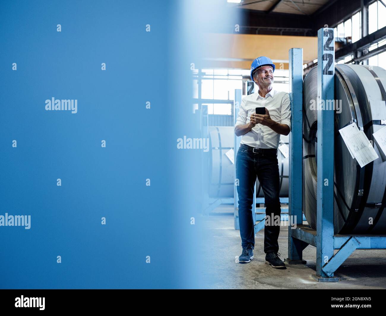 Lächelnder männlicher Ingenieur mit Mobiltelefon, das neben aufgerolltem Metallblech steht Stockfoto