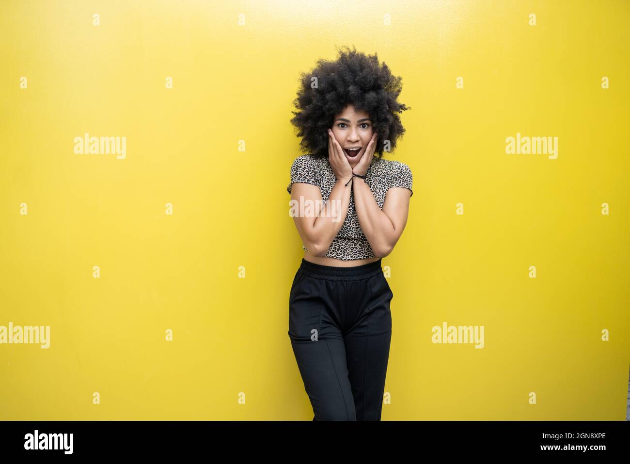 Überraschte Frau mit Händen am Kinn vor der gelben Wand Stockfoto