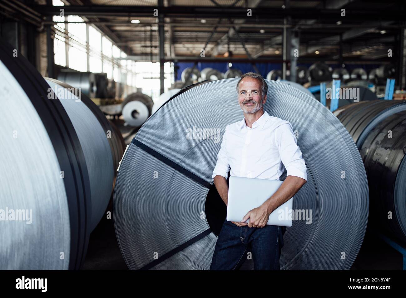 Lächelnder männlicher Geschäftsexperte mit Laptop, der in der Stahlindustrie steht Stockfoto