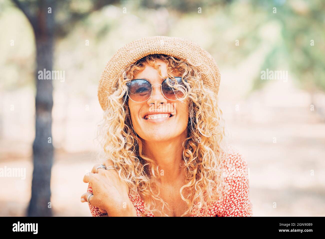 Lächelnde reife Frau mit lockigen blonden Haaren, die Sonnenbrille und Hut tragen Stockfoto