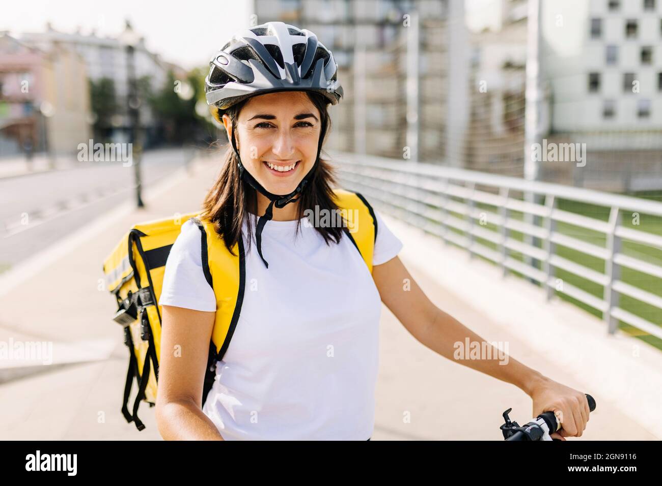 Lächelnde Frau mit essentieller Bedienung, die am sonnigen Tag Fahrradhelm und Lieferrucksack trägt Stockfoto