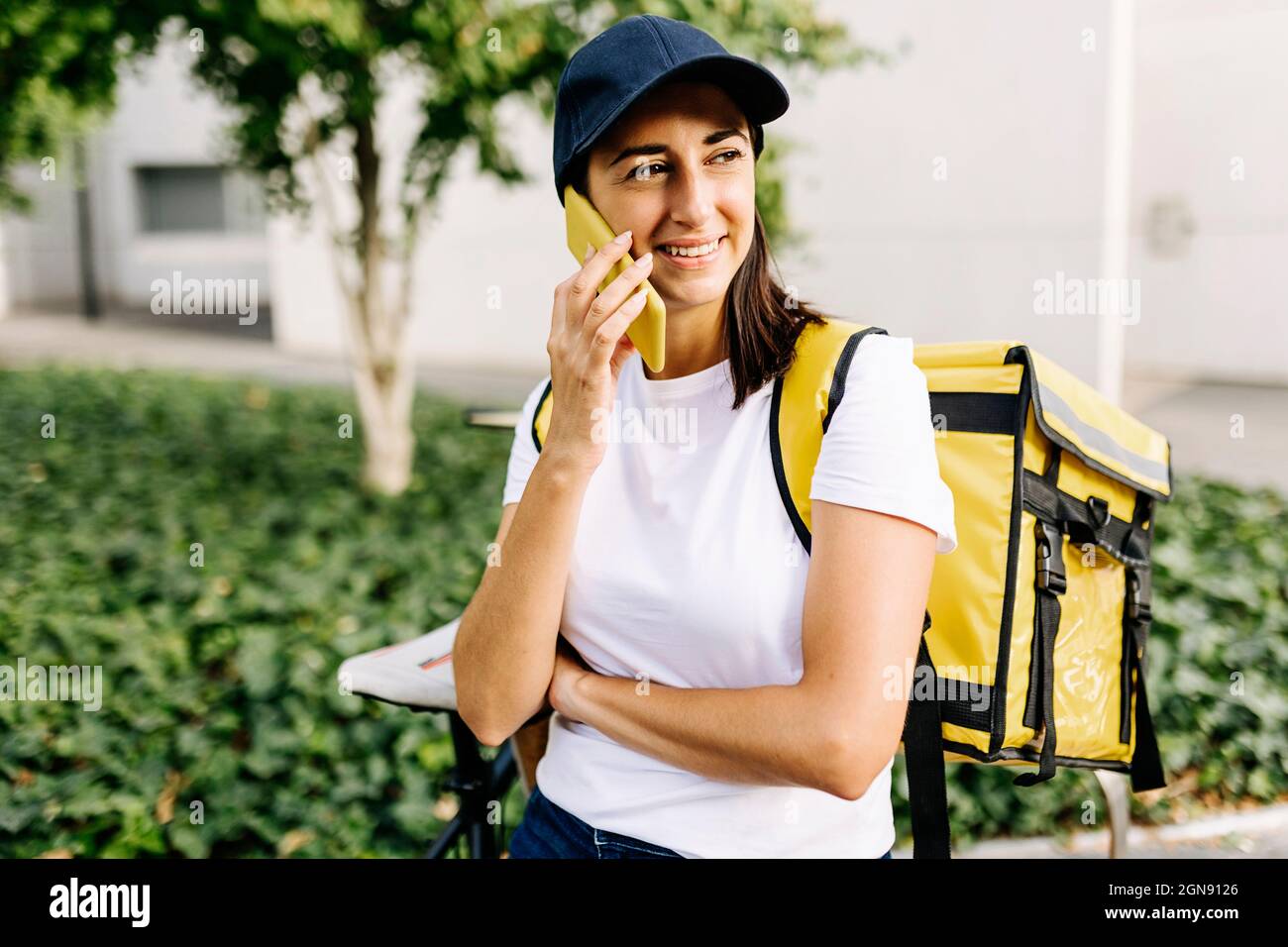 Lächelnde, unverzichtbare Servicefrau mit Rucksack für die Lieferung, die auf dem Mobiltelefon spricht Stockfoto