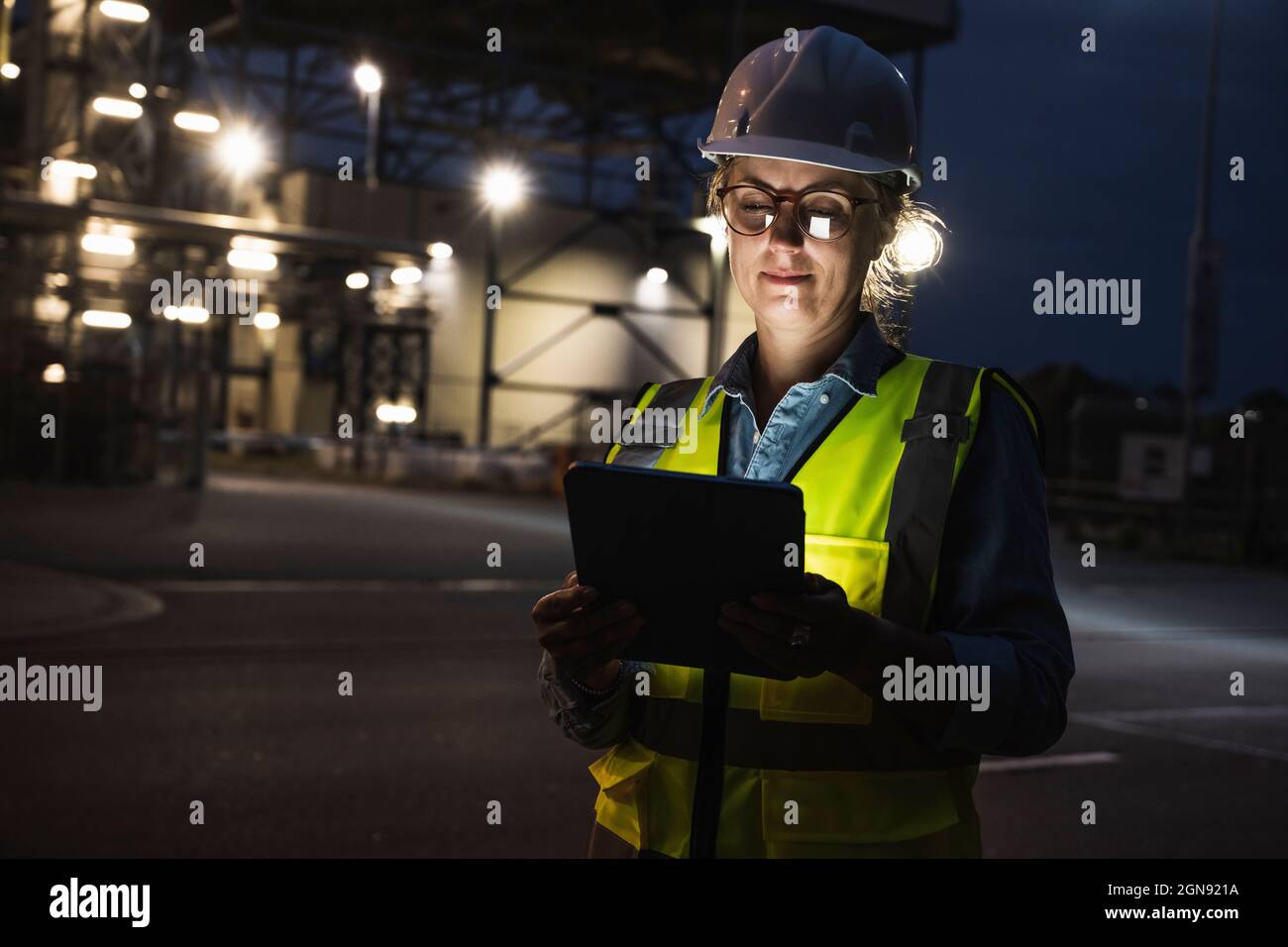 Ingenieurin mit Hardhut und digitalem Tablet außerhalb der Fabrik in der Nacht Stockfoto