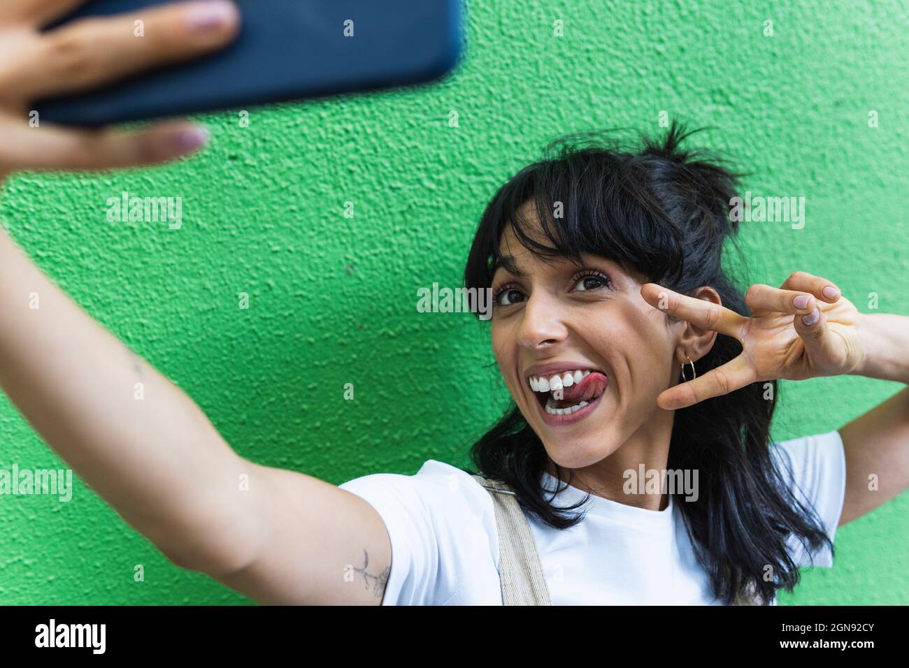 Fröhliche Frau, die beim Selfie ein Friedenszeichen zeigt und die Zunge heraussticht Stockfoto