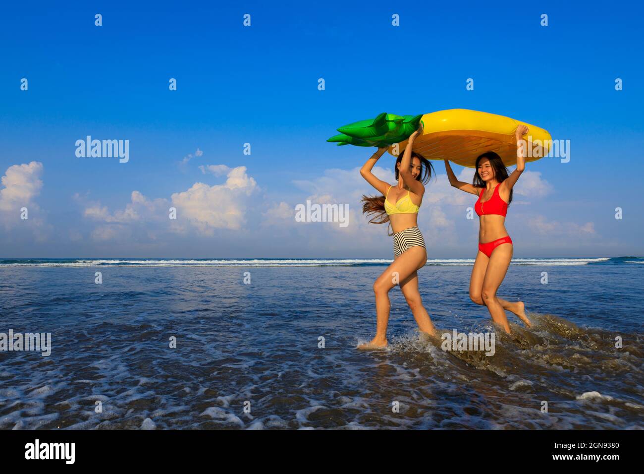 Fröhliche Freundinnen, die Ananas tragen, schweben auf dem Kopf, während sie im Urlaub am Strand genießen Stockfoto