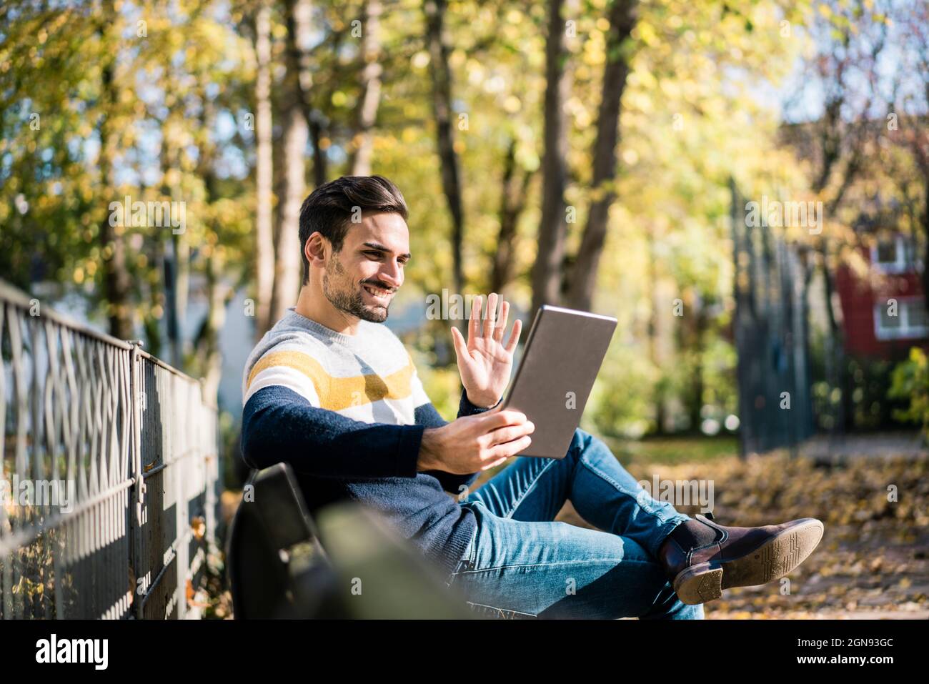 Junger Mann winkt bei einem Videoanruf über ein digitales Tablet im Park während eines sonnigen Tages mit der Hand Stockfoto