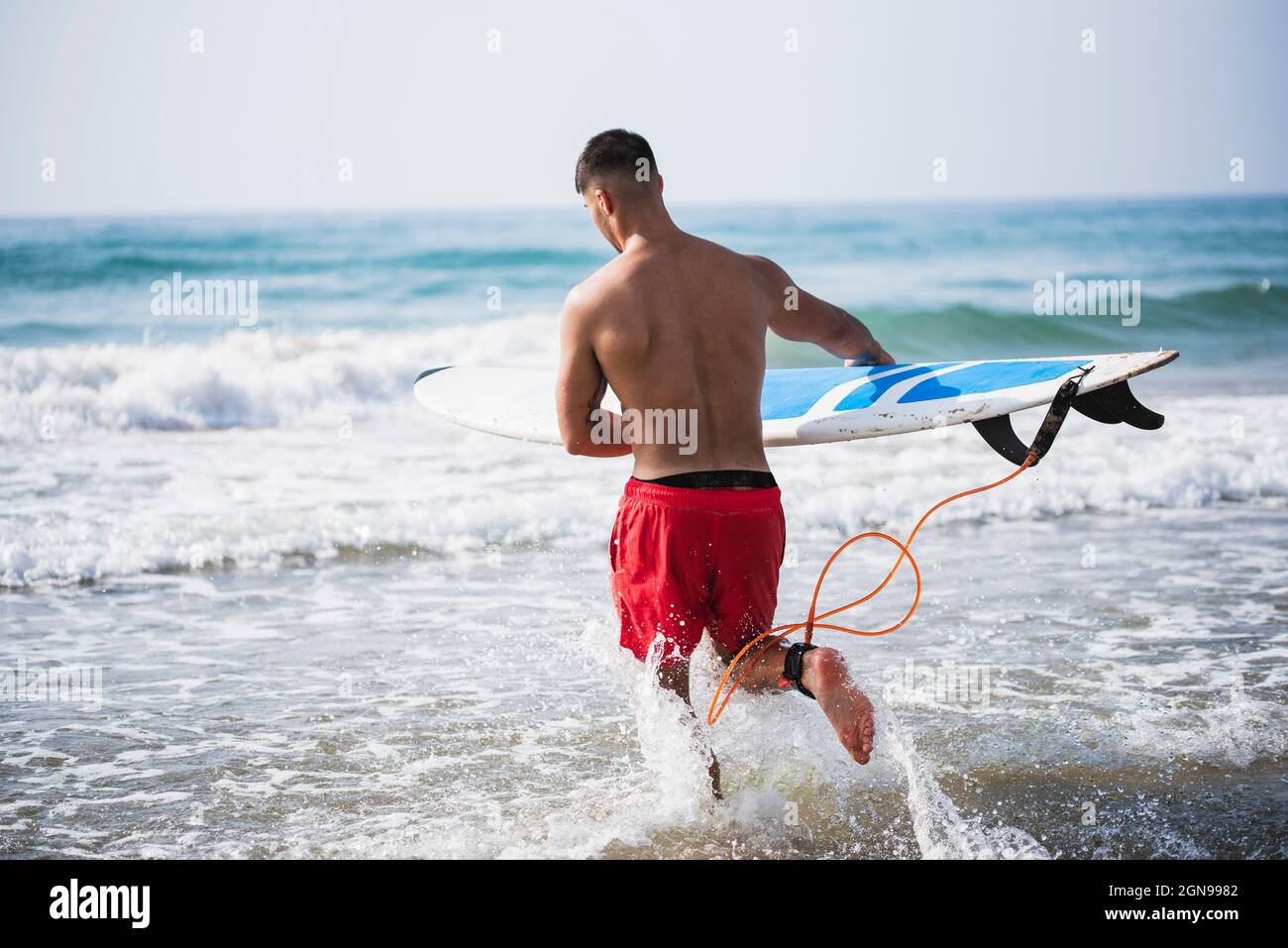 Rückansicht eines jungen Mannes, der ins Wasser läuft, um zu surfen. Stockfoto