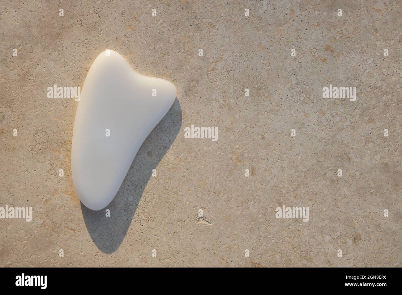 Gua Sha Scraper, weißes Achat-Massagegerät auf leicht strukturierter Oberfläche, Sonnenuntergangslicht, hartem Schatten. Platz zum Kopieren, flach liegend. Konzept Schönheit, Selbstpflege, Gesichtskörper Stockfoto