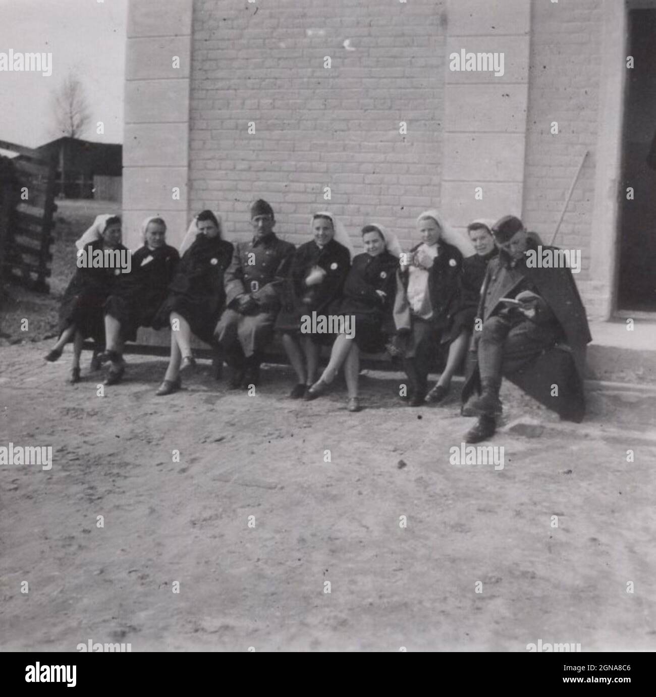 Seltener historischer Moment aus dem Zweiten Weltkrieg. Zwei Soldaten sitzen unter Krankenschwestern auf der Bank vor einem Haus oder einer Kirche. Die Krankenschwestern lächeln, essen, ruhen sich aus. Ein Soldat konzentriert sich auf den Fotografen, der andere gibt keinen Wurf, den er gerade liest. Zeitraum des 2. Weltkrieges. Quelle: Originalfoto (militärisches Subject) Stockfoto