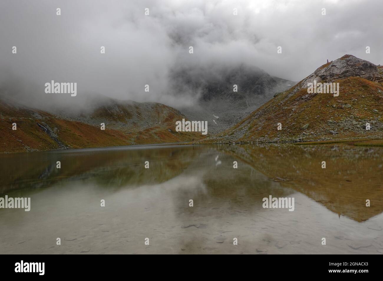Bergsee im Herbst unter niedrigen Wolken, Vegetation in Herbstfarben, Berge spiegeln sich in spiegelglattem Wasser Stockfoto