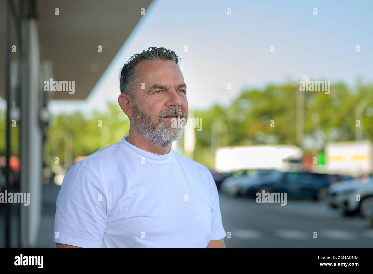 Nachdenklicher bärtiger älterer Mann, der auf einem Außenparkplatz steht und mit einem ruhigen nachdenklichen Ausdruck in einem Kopf- und Schulterporträt zur Seite blickt Stockfoto