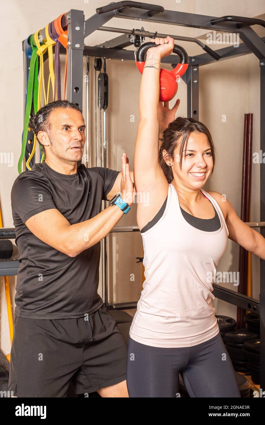 Frau tut Schulterpresse mit Kettlebell mit Hilfe des Trainers im Health Club Stockfoto