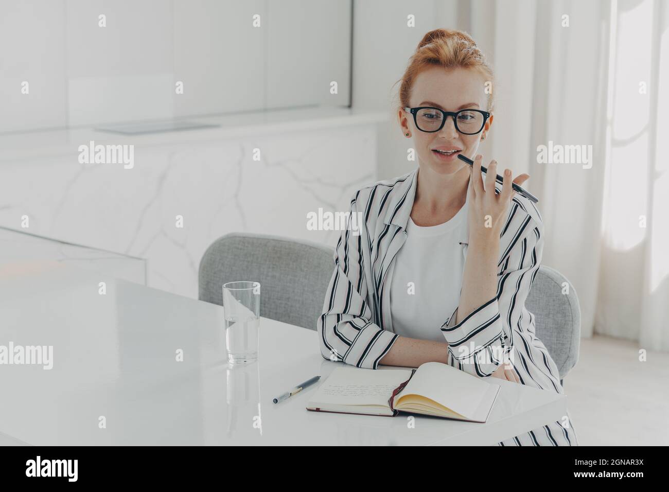 Foto von Rotschopf junge Frau nimmt Sprachanruf über Smartphone-Mikrofon sitzt am Desktop Stockfoto