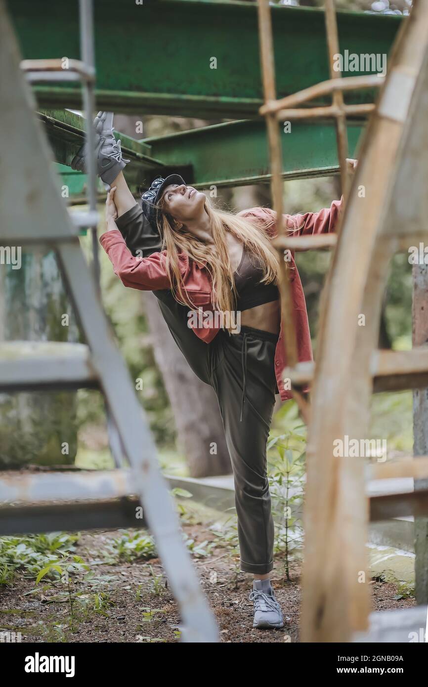 Eine Frau in legerem Outfit, die an einem Sommertag im Freien eine Dehnung macht, wobei ihr Bein über die Stange einer Metallstruktur geworfen wird Stockfoto