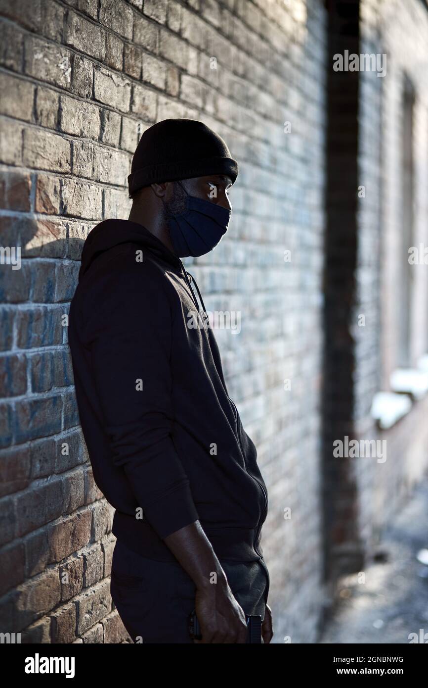 Junger afroamerikanischer Mann in schwarzer Maske, der auf der Straße steht und auf ein Opfer wartet, das ein Verbrechen begangen hat. Schwarzer afrikanischer Kerl in Hut und schwarzer Kleidung. Grausamer Mann o Stockfoto