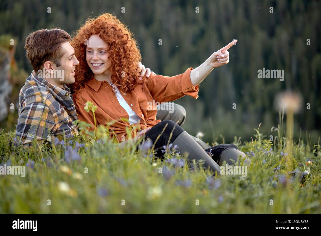 Lächelndes Paar, das in den Sommerbergen auf dem Land wandert. Glückliche Frau und Mann sitzen auf Gras. Weibliche und männliche Touristen in Freizeitkleidung erkunden die Natur, Stockfoto
