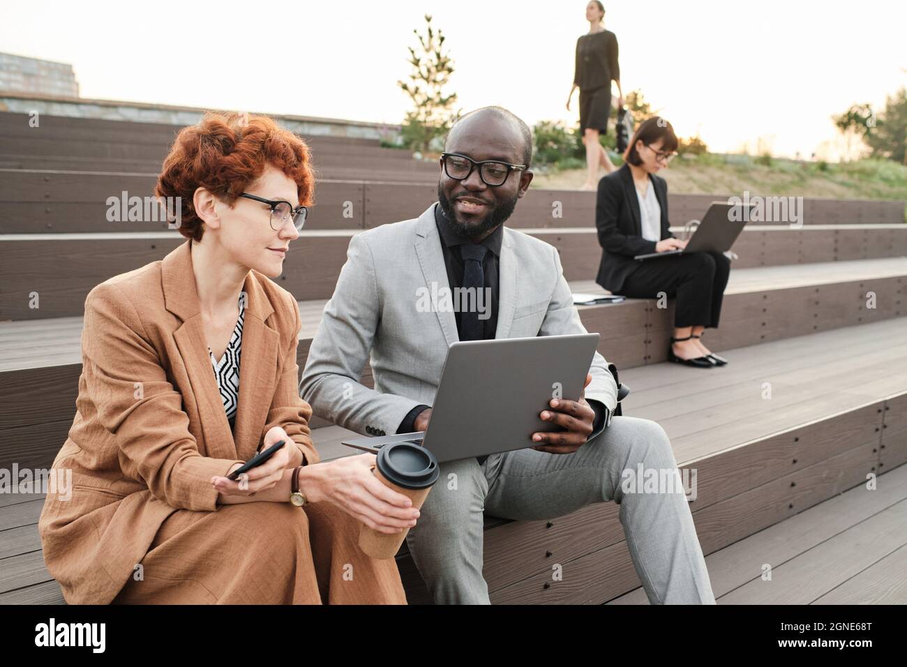 Horizontales Porträt eines modernen afroamerikanischen Mannes, der im Freien sitzt, während sein Kollege ihr etwas auf seinem Laptop vorführt Stockfoto