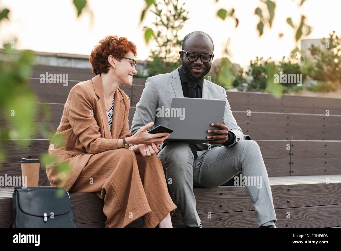 Horizontale, mittellange Aufnahme eines erfolgreichen afroamerikanischen Mannes und einer kaukasischen Frau, die auf einer Bank im Stadtpark sitzen und Daten auf einem Laptop betrachten Stockfoto