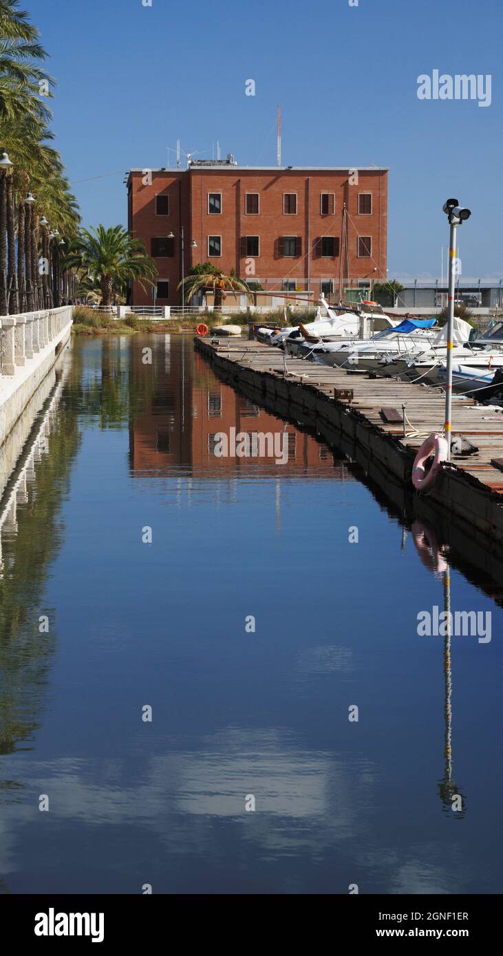 Im Hafen von Olbia spiegelt sich ein rechteckig gefliestes Gebäude in das Meerwasser und bildet neben den geschleppten Grafikbooten eine quadratische Box. Sardinien, Italien Stockfoto