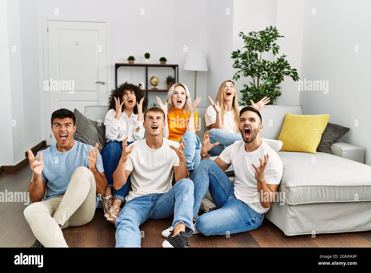 Gruppe von Menschen, die zu Hause auf dem Sofa und dem Boden sitzen, sind verrückt und brüllen mit aggressivem Ausdruck und erhobenen Armen Stockfoto