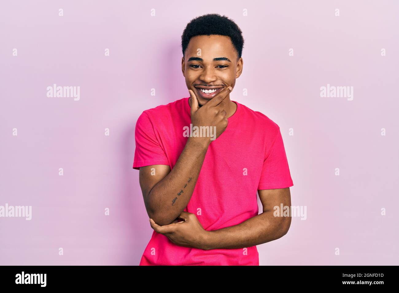 Junger afroamerikanischer Mann mit Kasua-T-Shirt, der mit gekreuzten Armen und erhobener Hand auf dem Kinn lächelnd zuversichtlich auf die Kamera blickt. Stockfoto