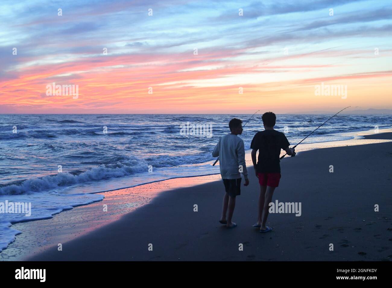 Rückansicht von zwei Jungen im Teenageralter, die bei Sonnenuntergang am Ufer des Sandstrandes mit Angelruten spazieren, Marina di Castagneto Carducci, Toskana Stockfoto