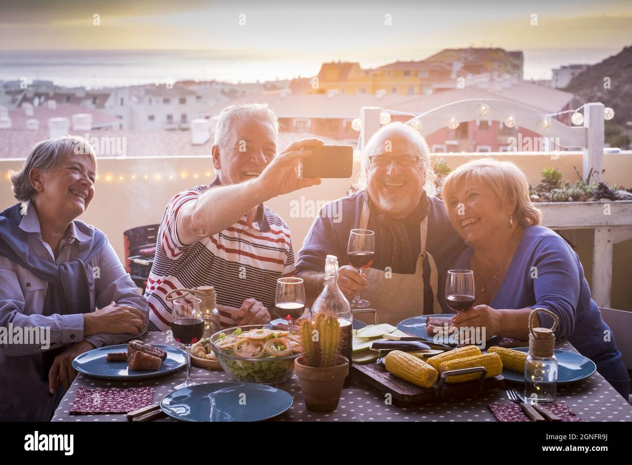 Zwei fröhliche ältere Paare, die auf der Terrassenparty speisen und Selfie mit dem Smartphone machen. Ältere Menschen haben Spaß beim Essen auf dem Dach. Alte Familie Stockfoto