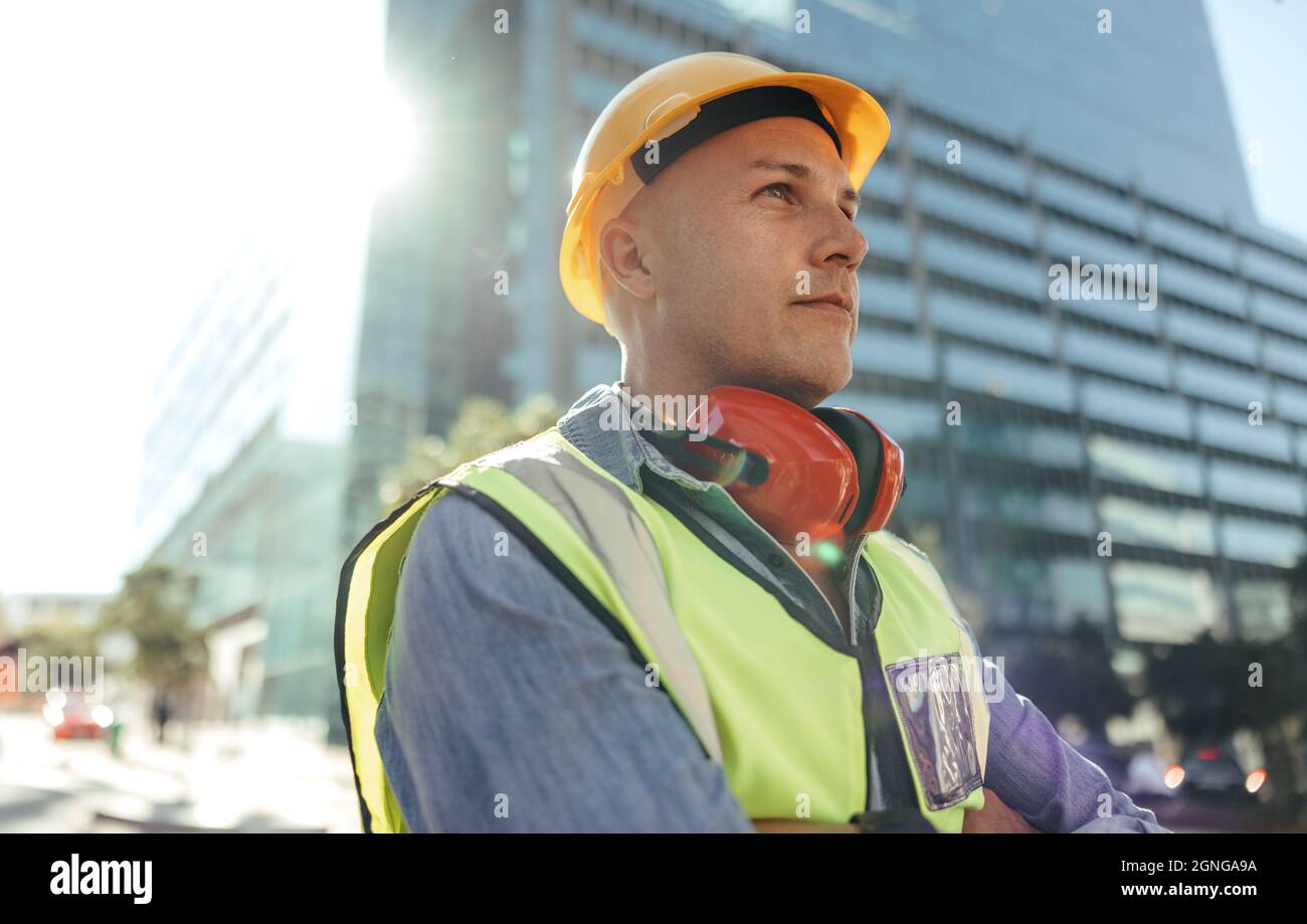 Arbeiter mit blauem Kragen schaut nachdenklich weg, während er mit gekreuzten Armen in der Stadt steht. Bauarbeiter mit mittlerem Erwachsenenalter, der vor der hohen Tür steht Stockfoto