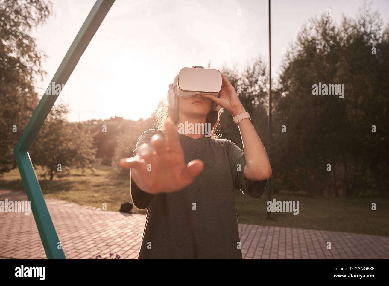 Nahaufnahme eines Mädchens, das mit den Händen eine Virtual-Reality-Brille auf dem Kopf hält und auf einem Hintergrund von grünen Bäumen steht. Er reicht eine Hand an die Kamera. Hochwertige Fotos Stockfoto