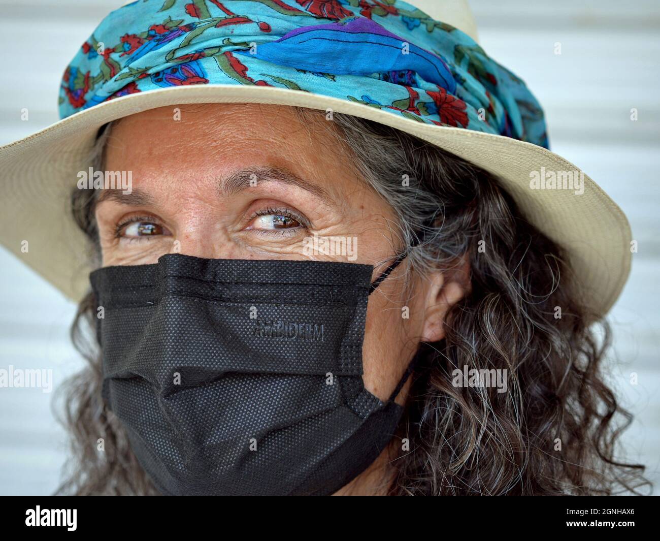 Charmante positive reife kaukasische Frau mit schönen braunen Augen trägt panama-Hut mit stilvollen blauen Hutband und schwarze Einweg-Gesichtsmaske. Stockfoto