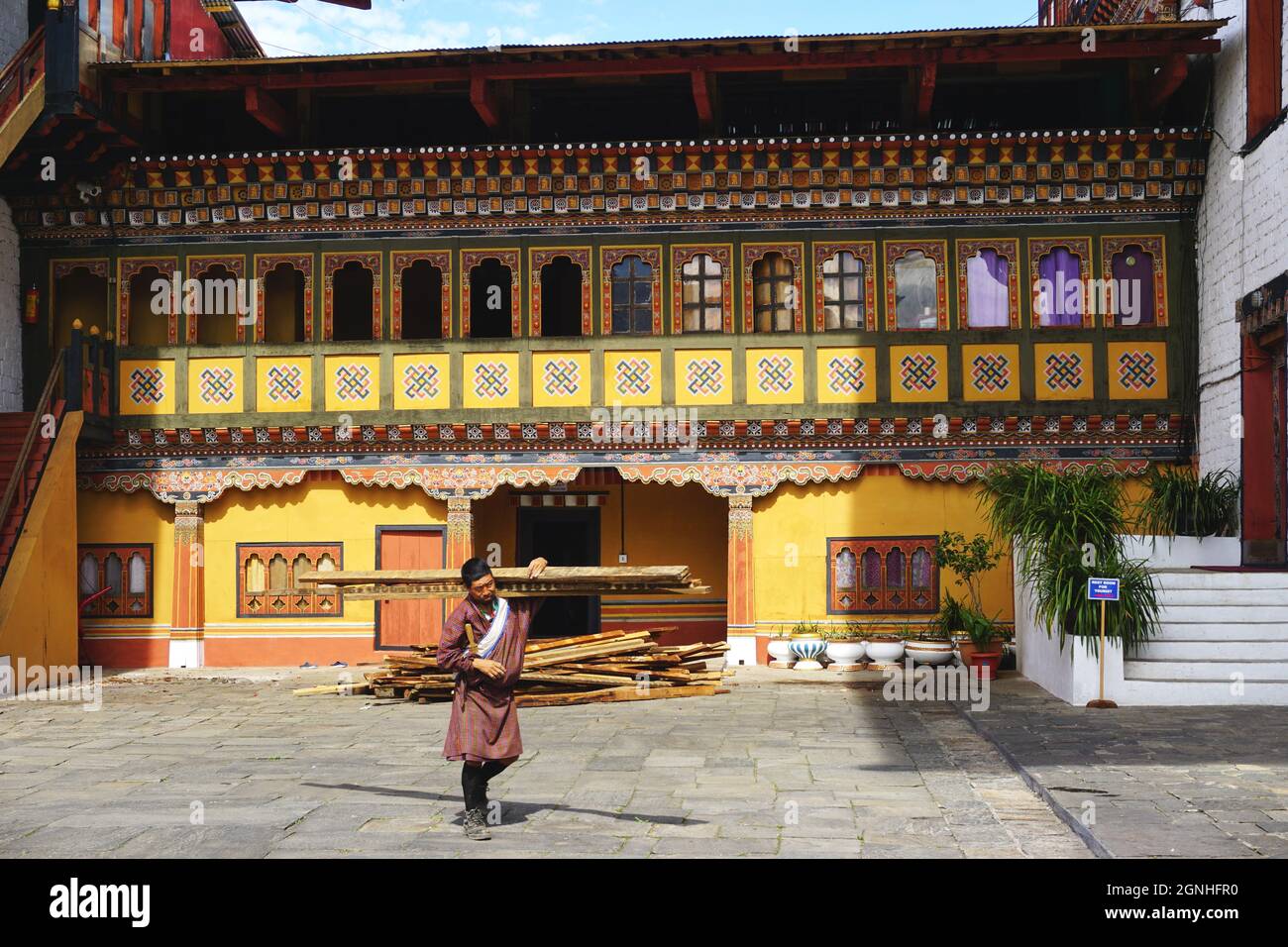 Ein Arbeiter, der eine traditionelle bhutanische gho trägt, trägt Holz an einem bunt bemalten Gebäude auf dem Gelände des Tashichho Dzong in Thimphu, Bhutan vorbei. Stockfoto