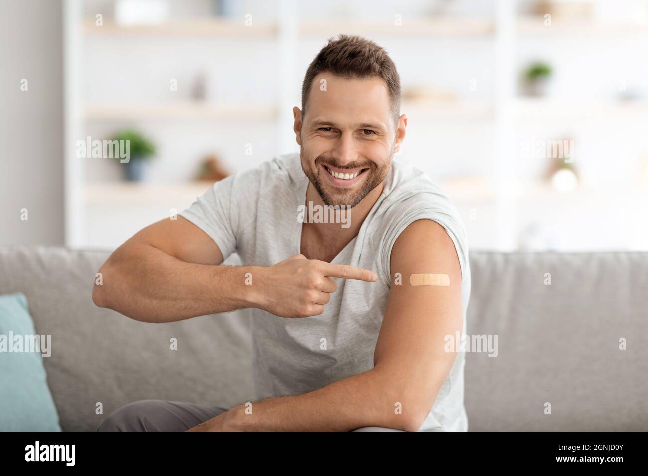 Der junge geimpfte Kerl zeigt mit medizinischem Gips auf die Schulter und lächelt weithin und fühlt sich nach der Impfung gesund Stockfoto