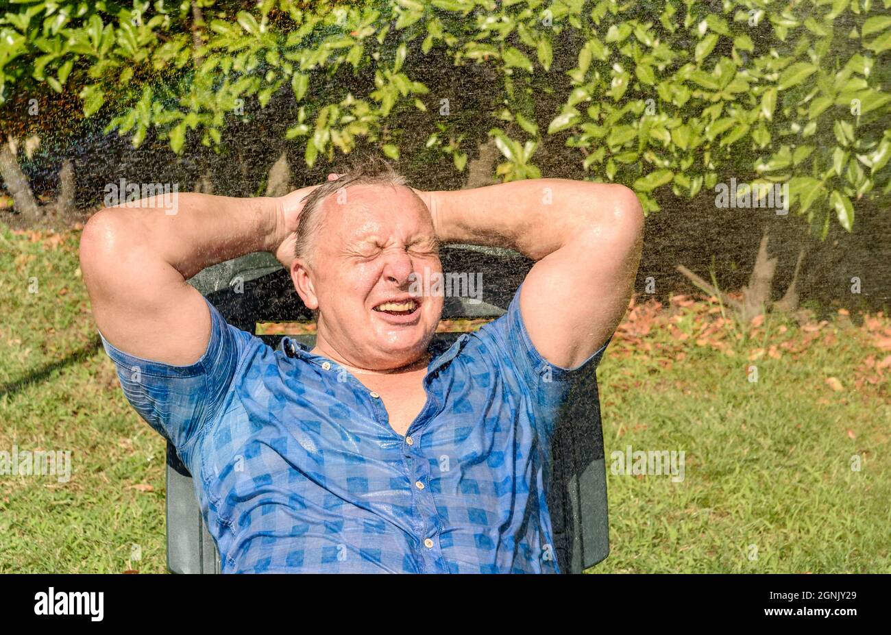 Porträt eines glücklichen reifen Mannes unter spritzendem Wasser an einem heißen Tag im Garten. Stockfoto