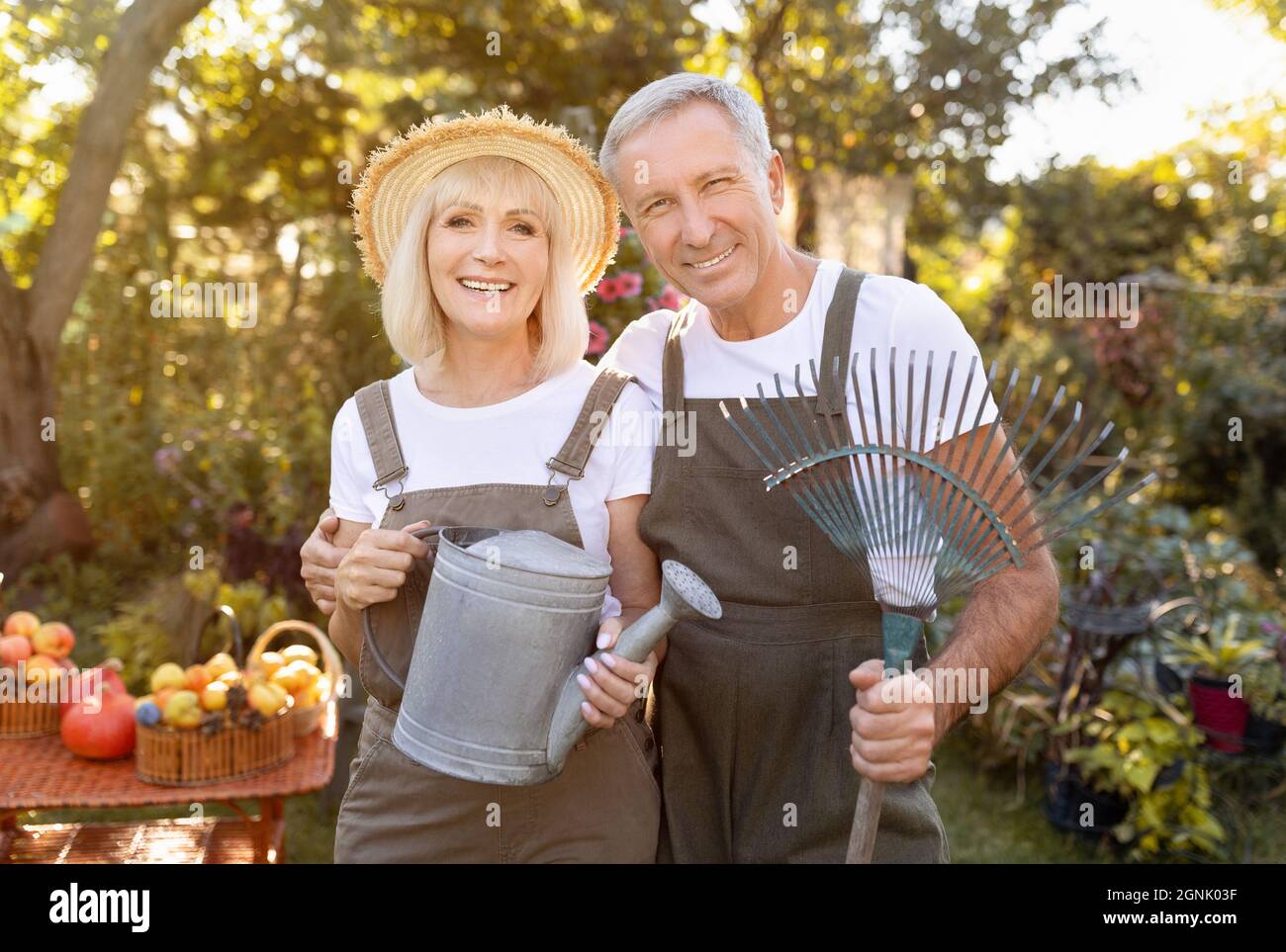 Schöne ältere Ehepartner halten Gartengeräte, arbeiten im Garten an sonnigen Tagen, Frau mit Gießkanne, Mann mit Rechen Stockfoto