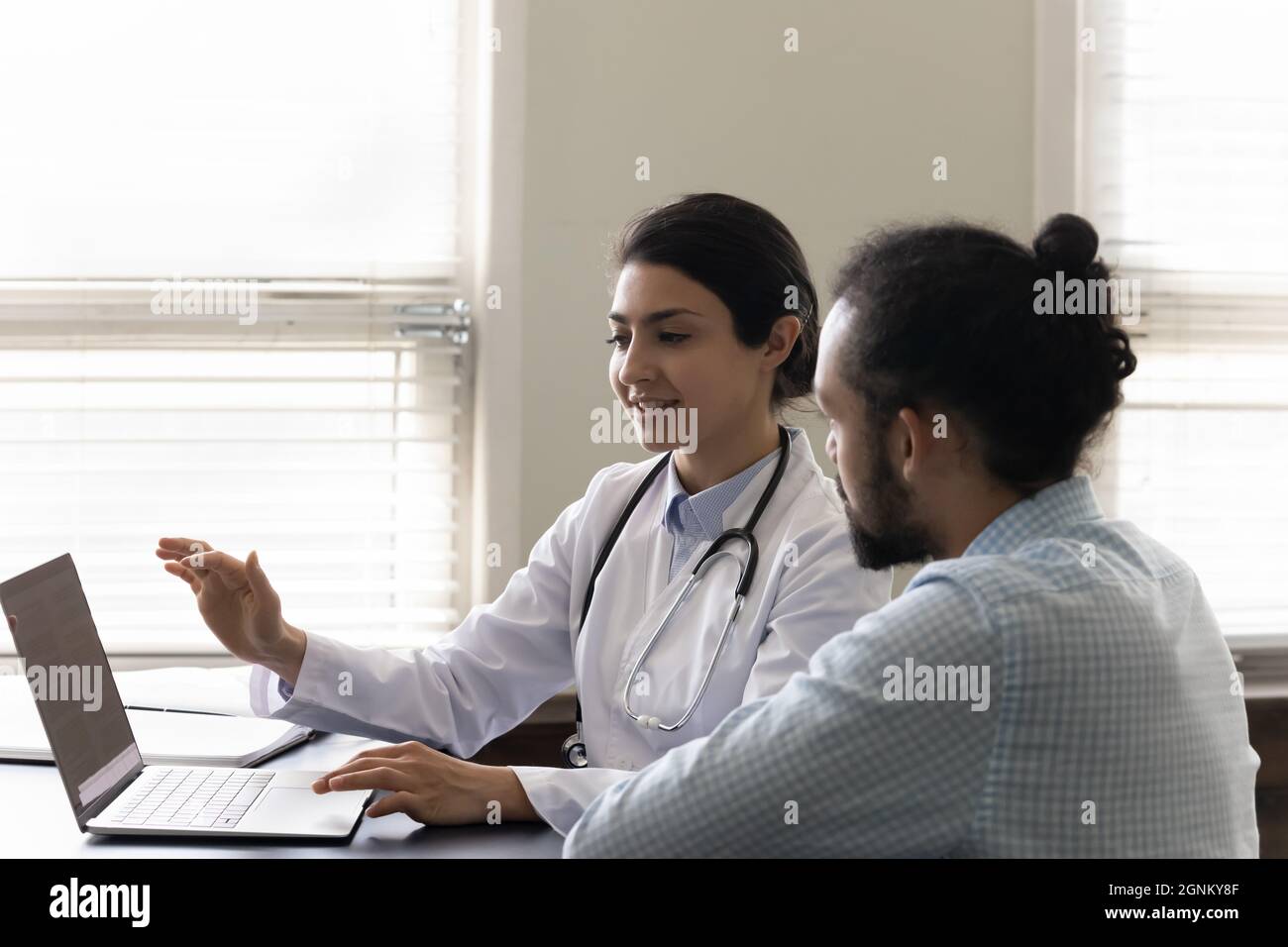 Lächelnder einfühlsamer indischer Praktizierender zeigt dem männlichen Patienten den Laptop-Bildschirm Stockfoto