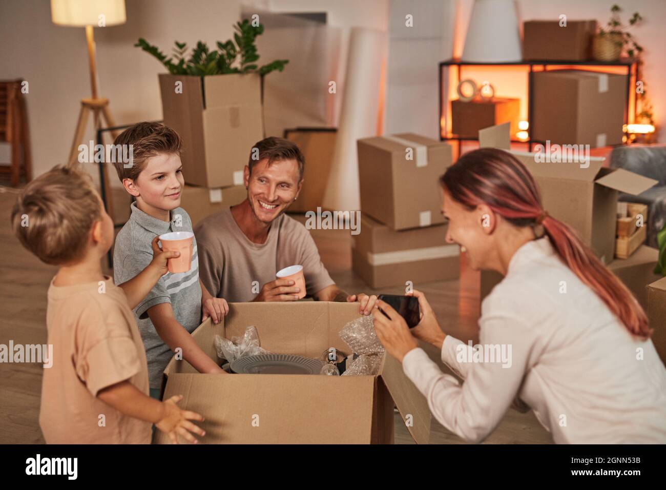 Porträt einer jungen glücklichen Familie, die Kisten mit Geschirr auspackt und Fotos macht, während sie in ein neues Zuhause einzieht, Platz kopiert Stockfoto